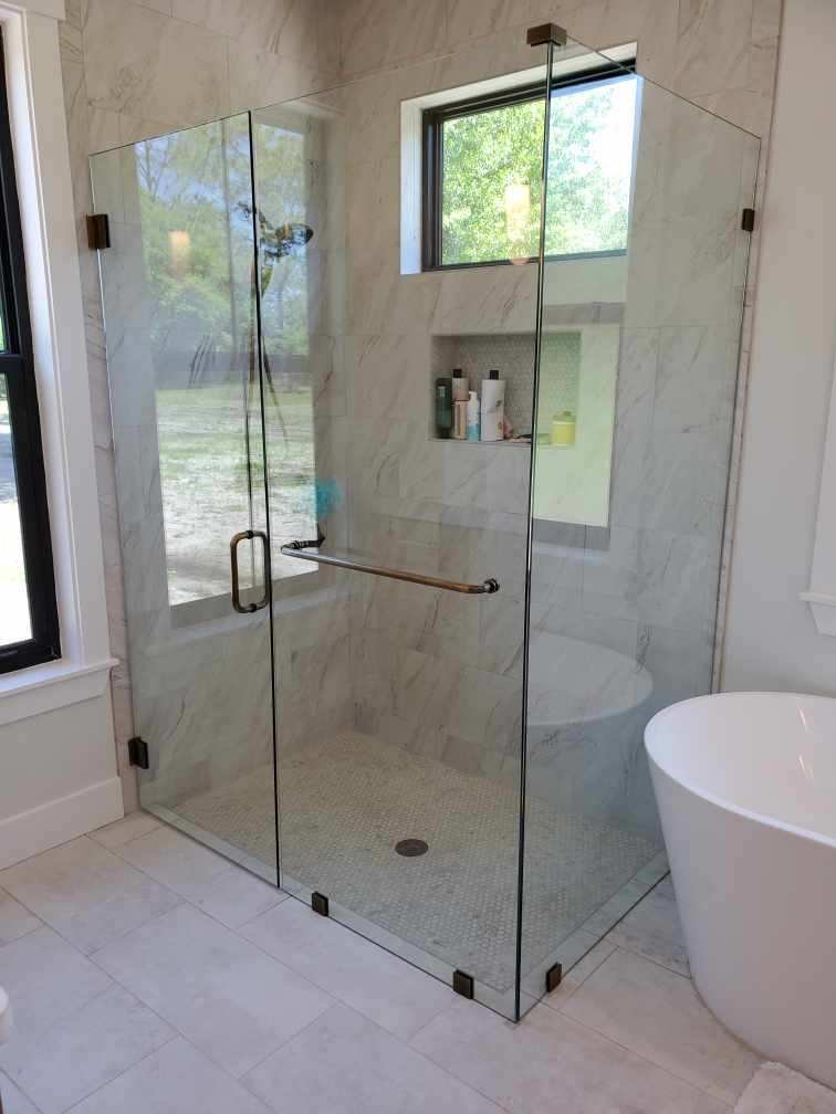 Corner glass shower enclosure with metal grab bar and tiled walls in a bathroom with nearby freestanding bathtub.