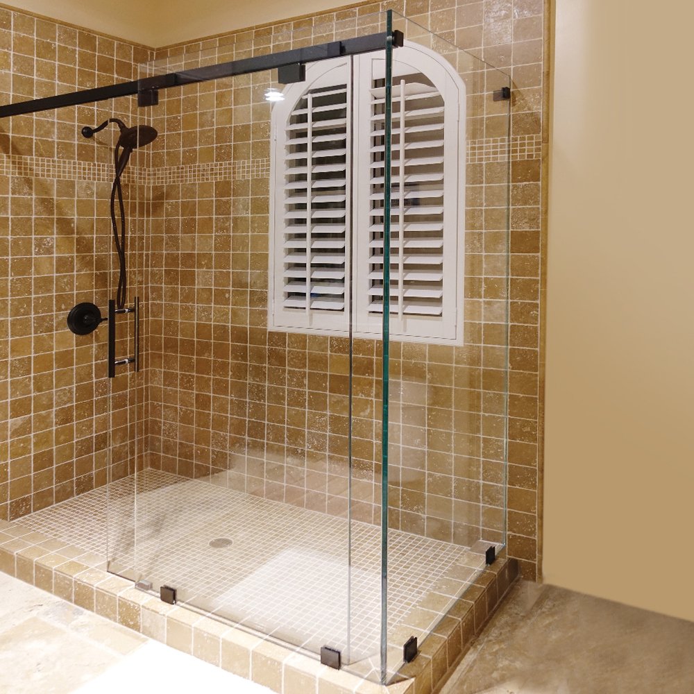 Sliding glass shower enclosure with dark hardware in a bathroom with brown tile walls and a white shuttered window.