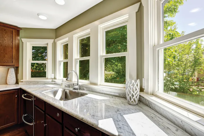 Multiple white windows installed along a marble kitchen counter top with dark wood cabinetry.